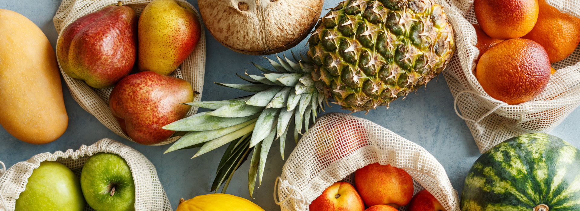 Verschiedenes Obst in Stoffbeuteln, darunter Ananas, Orangen, Äpfel und Birnen.
